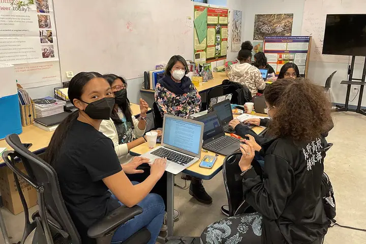 Students in masks work on laptops together around tables in a classroom.