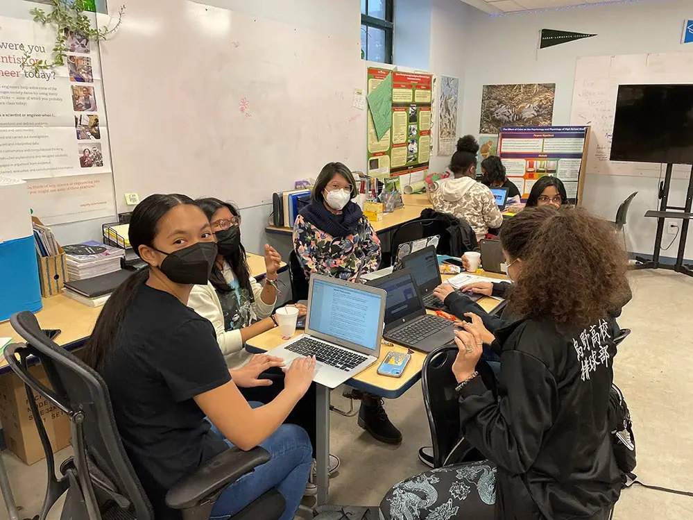 Students in masks work on laptops together around tables in a classroom.