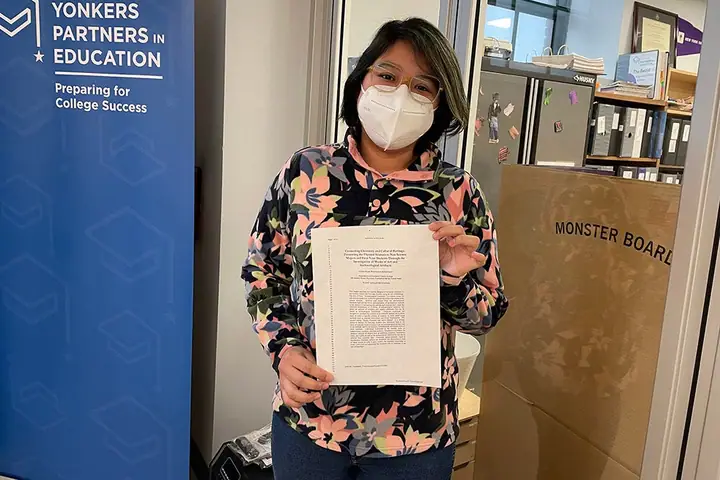 A student in a mask holds up a printed academic paper beside a Yonkers Partners in Education banner.