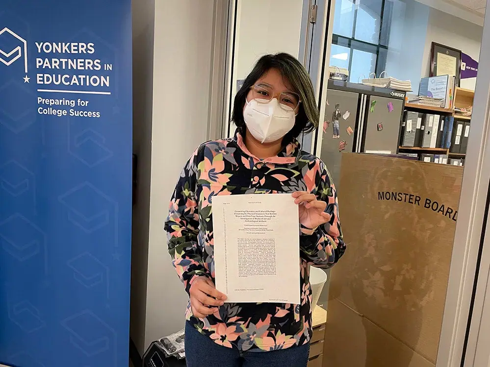 A student in a mask holds up a printed academic paper beside a Yonkers Partners in Education banner.