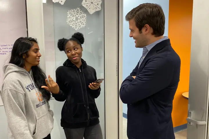 Two high school students talk with a young man in a blazer in a hallway hung with paper snowflakes.
