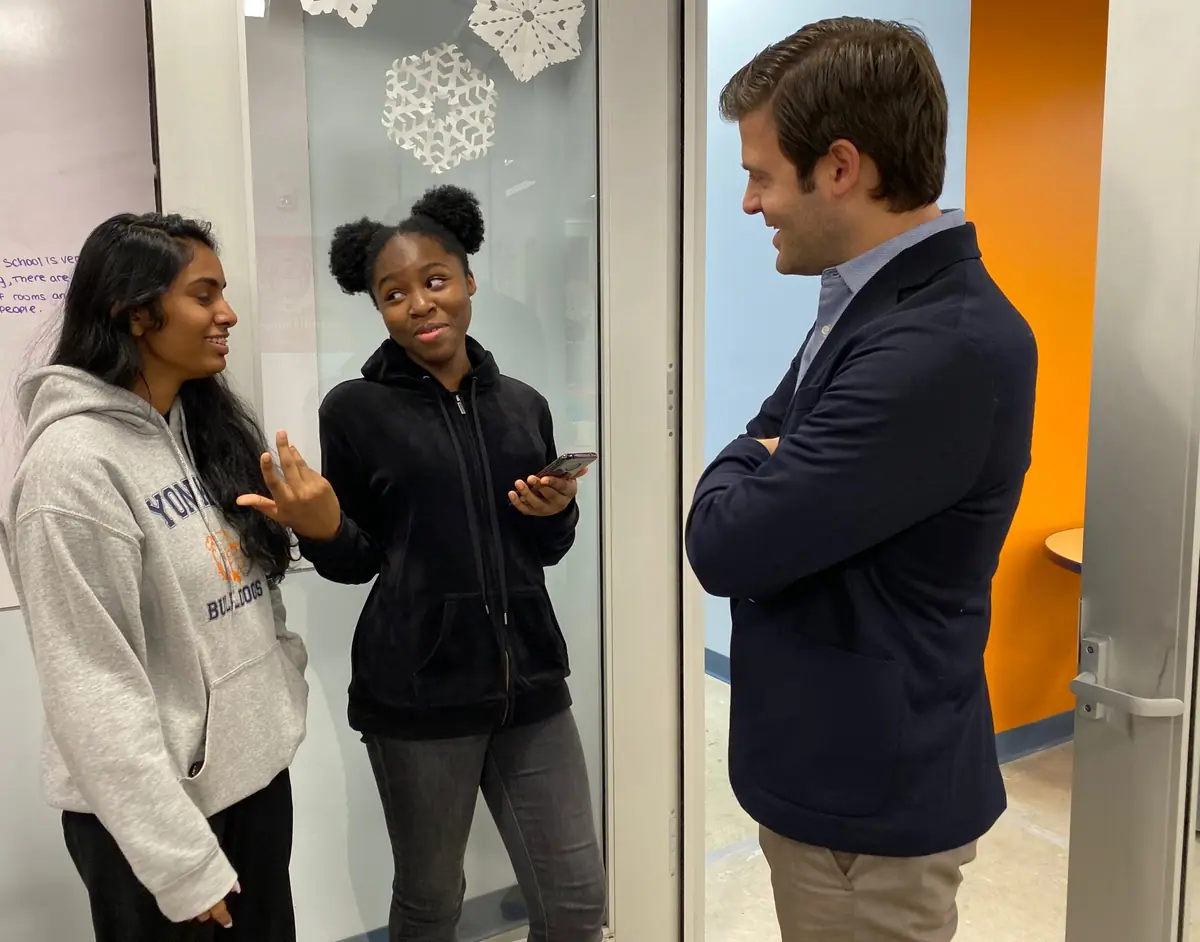 Two high school students talk with a young man in a blazer in a hallway hung with paper snowflakes.