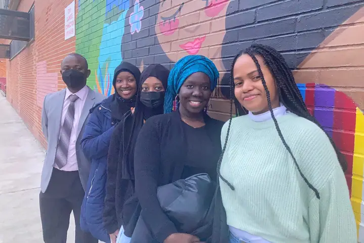 A man in a suit stands with four young women in front of a colorful mural on a brick wall.