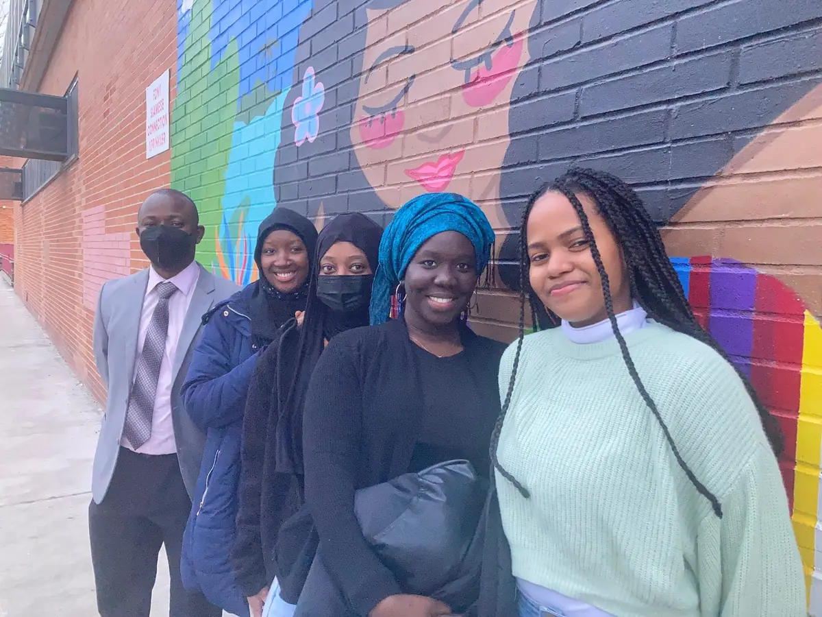 A man in a suit stands with four young women in front of a colorful mural on a brick wall.