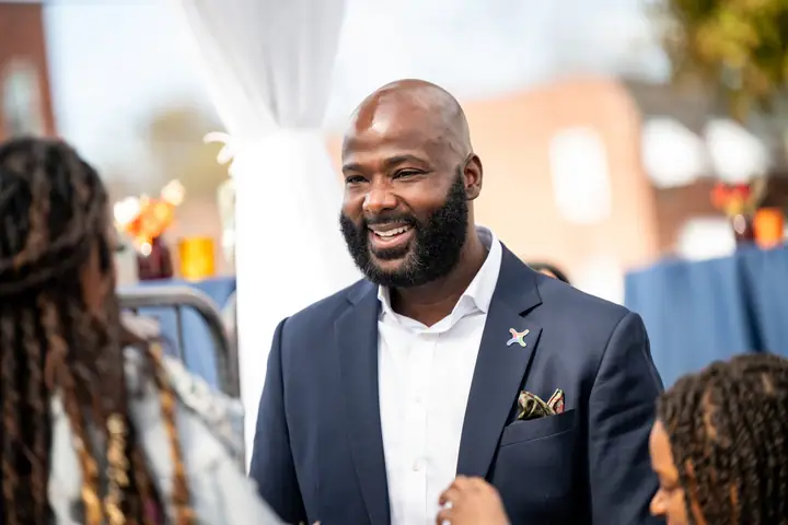 A smiling man in a blazer greets people at an outdoor gathering.
