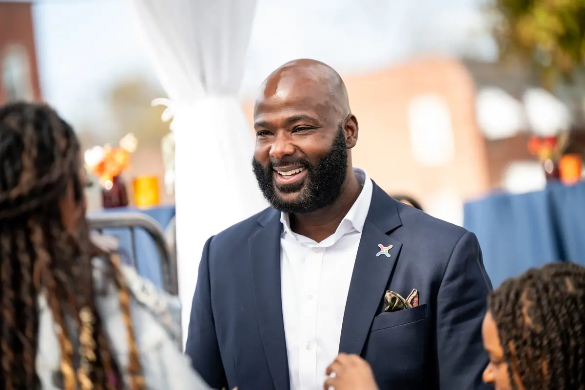 A smiling man in a blazer greets people at an outdoor gathering.