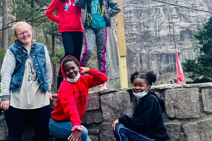 Girls and a woman pose on a stone wall below a granite mountain face.