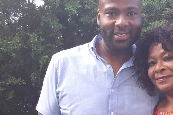 A man and an older woman smile cheek to cheek outdoors, in front of green trees.