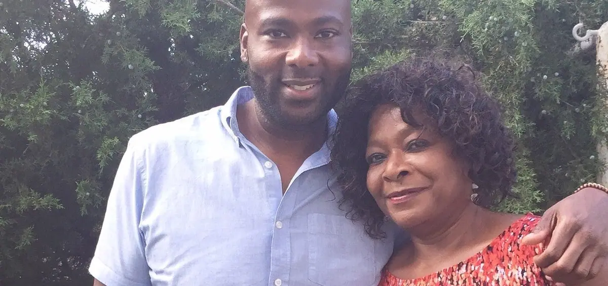 A man and an older woman smile cheek to cheek outdoors, in front of green trees.
