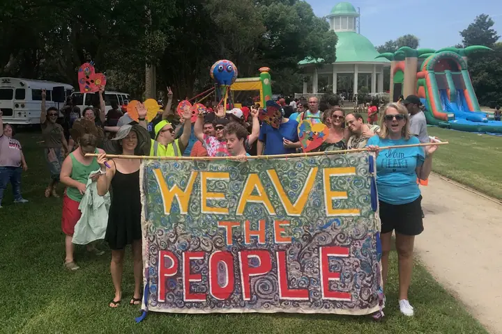 People in a park hold up a hand-painted WEAVE THE PEOPLE banner at an outdoor festival.