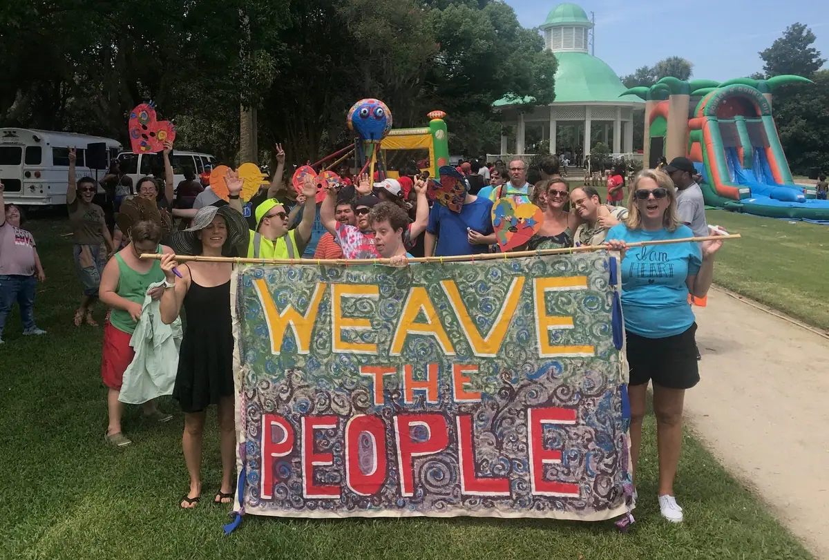 People in a park hold up a hand-painted WEAVE THE PEOPLE banner at an outdoor festival.