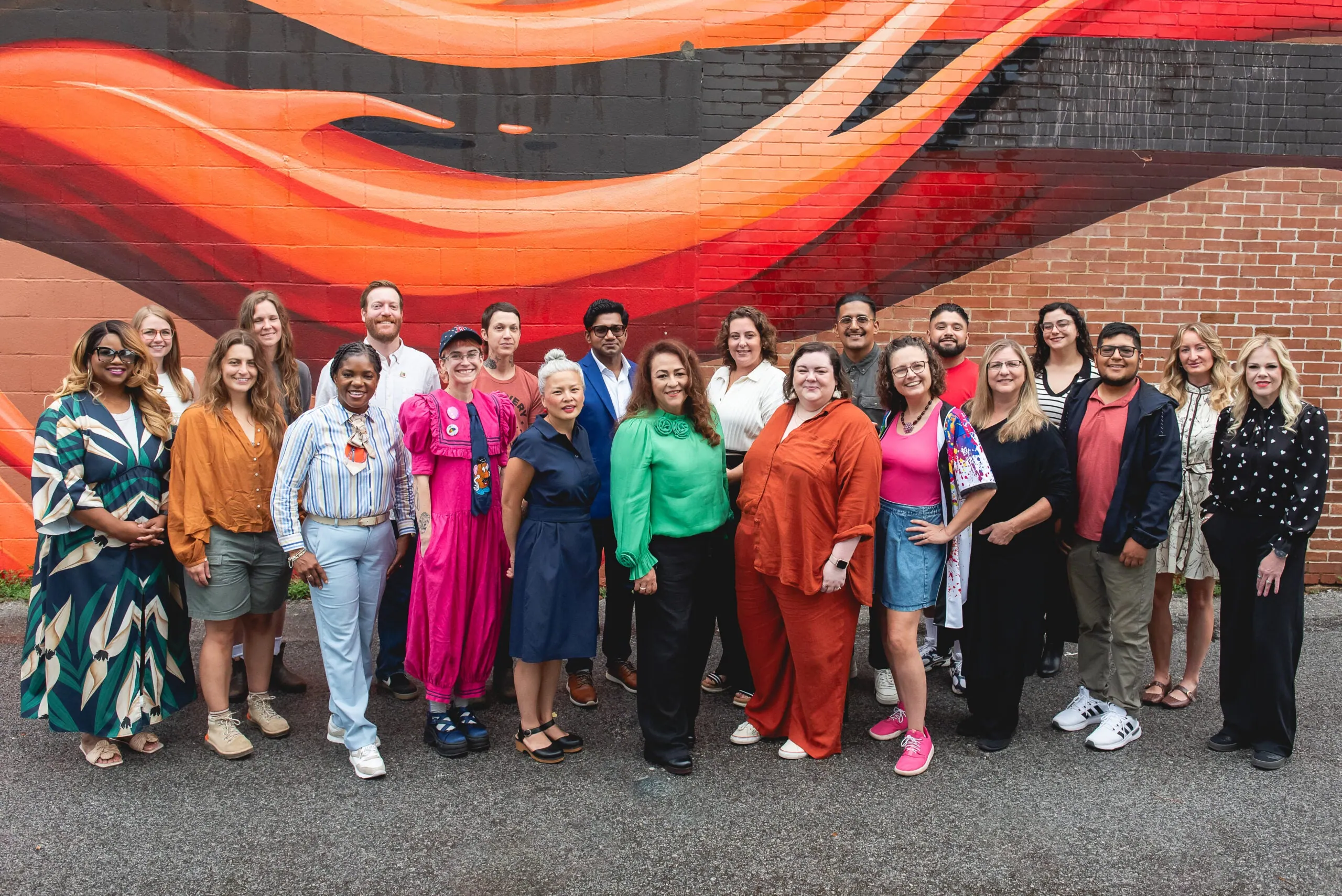 About two dozen people stand together in front of a painted orange and red mural on a brick wall.