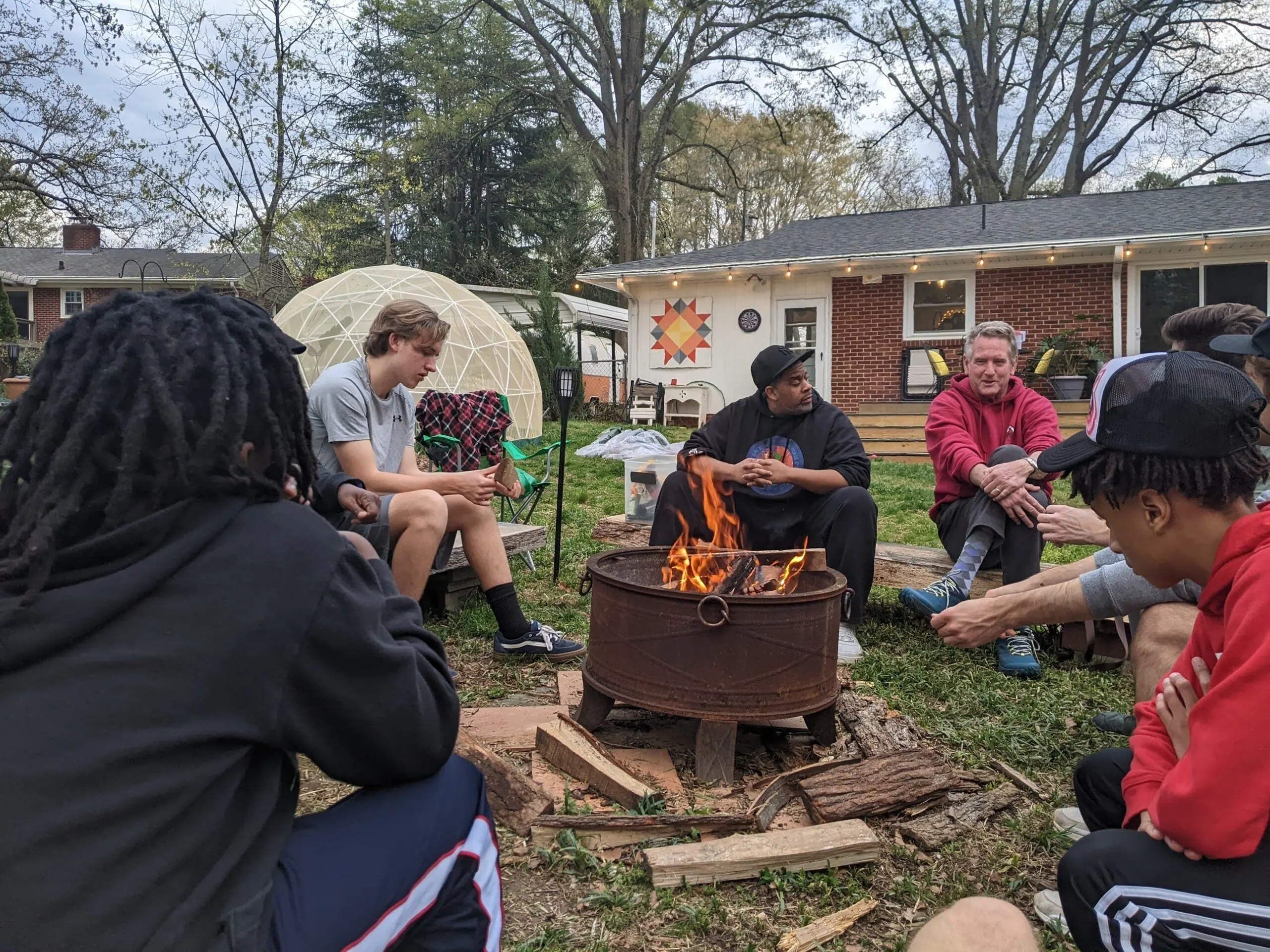 Teenage boys and adult mentors sit in a circle around a fire pit in a back yard.