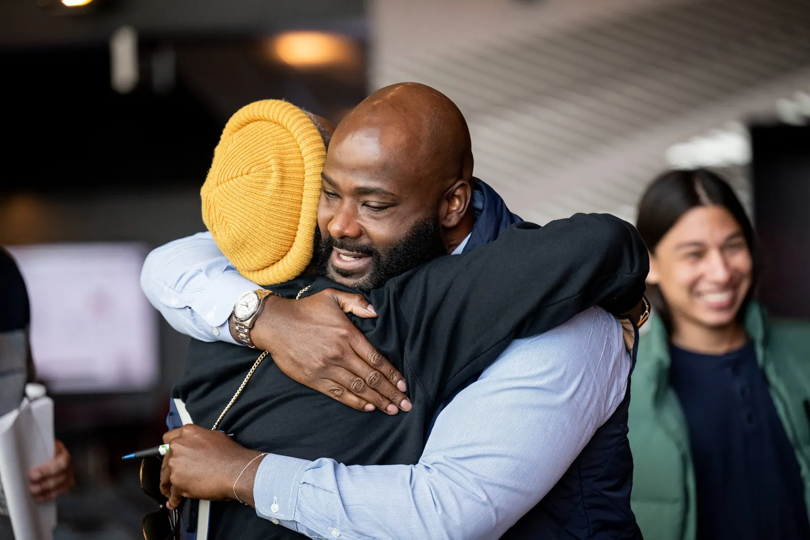 Frederick Riley embraces a guest in a yellow knit hat while a woman beside them smiles.