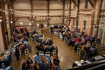 Guests at round tables fill the barn floor under strings of lights.