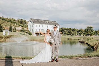 A bride and groom on the path by the pond, the barn on the hill behind them.